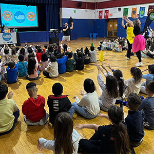 students sitting on gym floor watching adults with their hands up